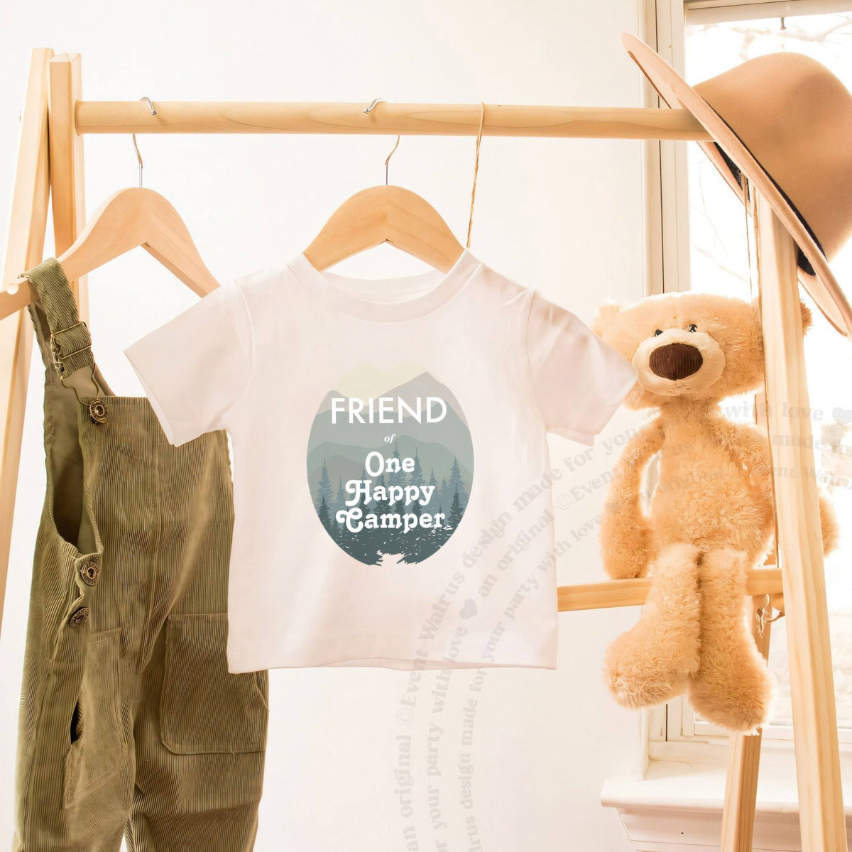 White Friend of One Happy Camper toddler tee on a wooden clothes rack beside a teddy bear.