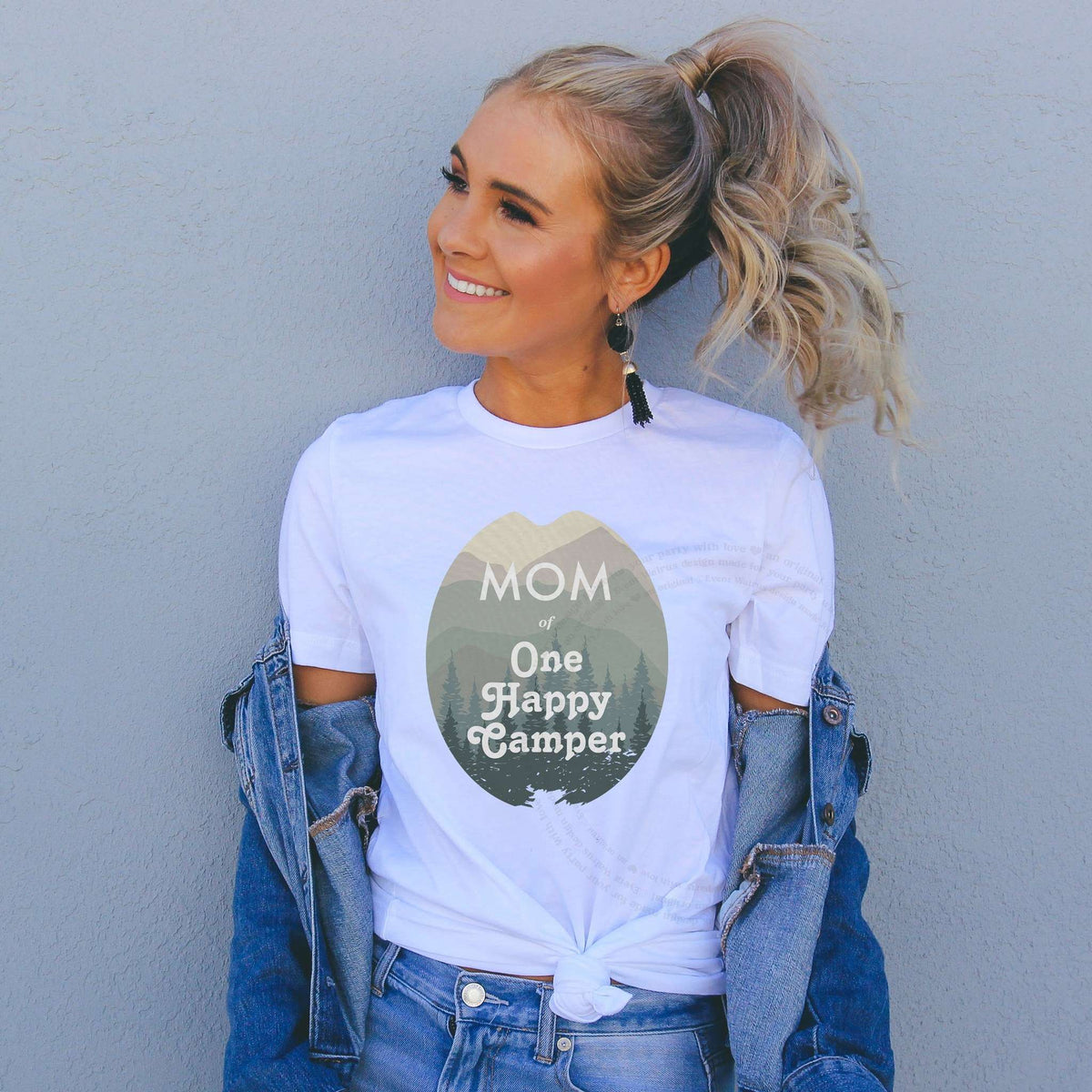 Smiling woman in a denim jacket wearing a white Mom of One Happy Camper shirt.