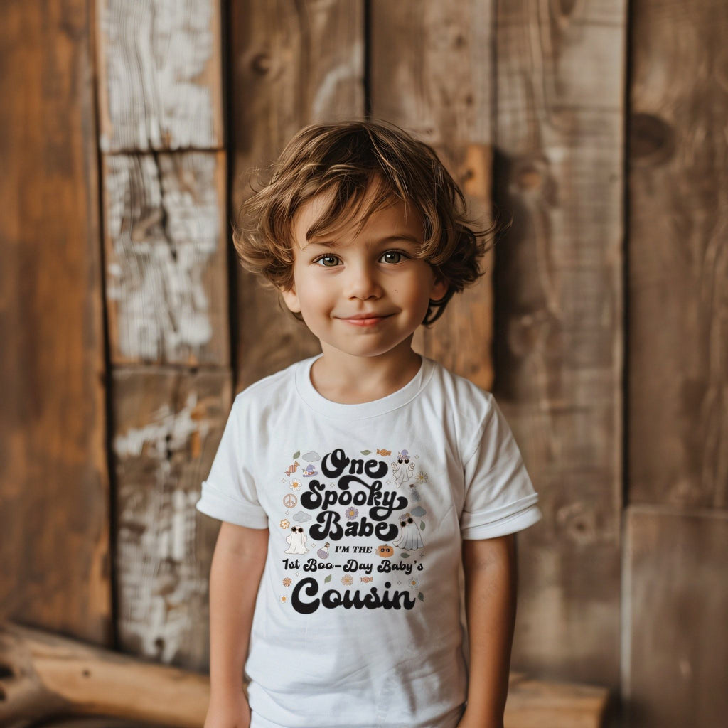 Toddler boy against a weathered wood wall in a white One Spooky Babe Cousin tee.
