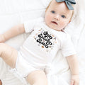 Baby girl with a gray bow lying in a white One Spooky Babe 1st Boo-Day bodysuit.