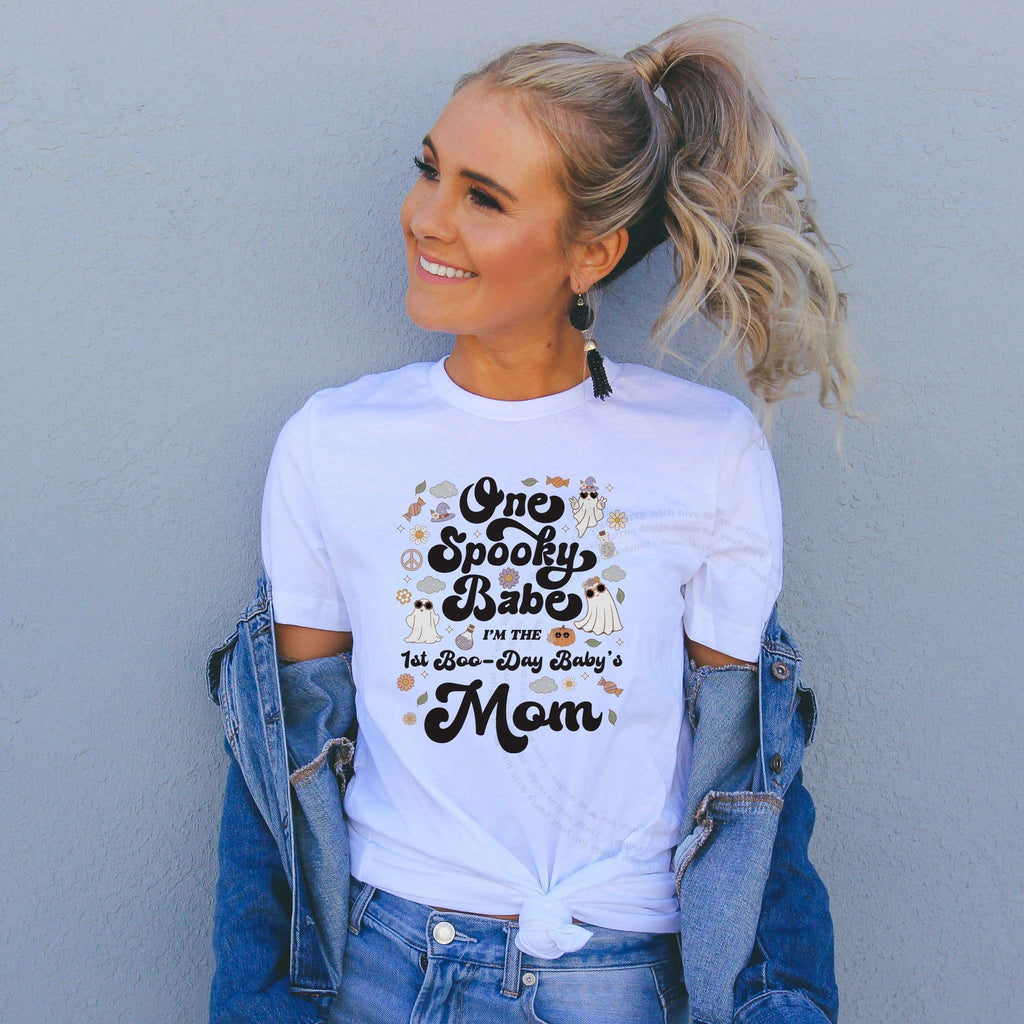 Smiling woman in a denim jacket wearing a white One Spooky Babe 1st Boo-Day Baby's Mom shirt.