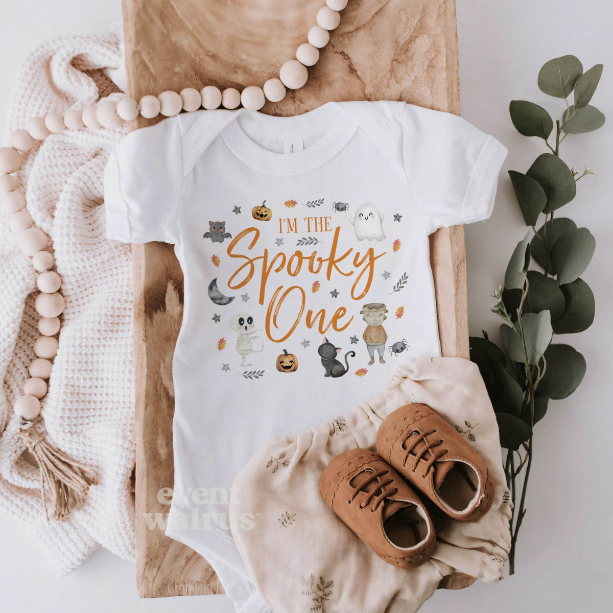 White I'm the Spooky One bodysuit laid flat with a knit blanket, wooden beads and baby shoes.