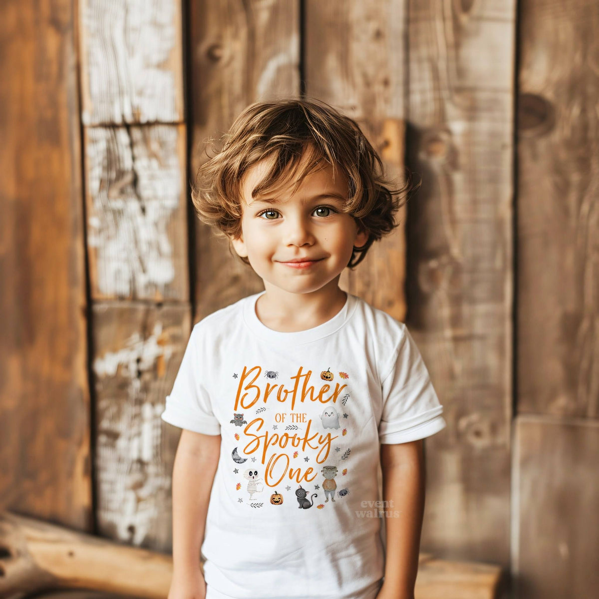 Toddler boy against a weathered wood wall in a white Brother of the Spooky One tee.