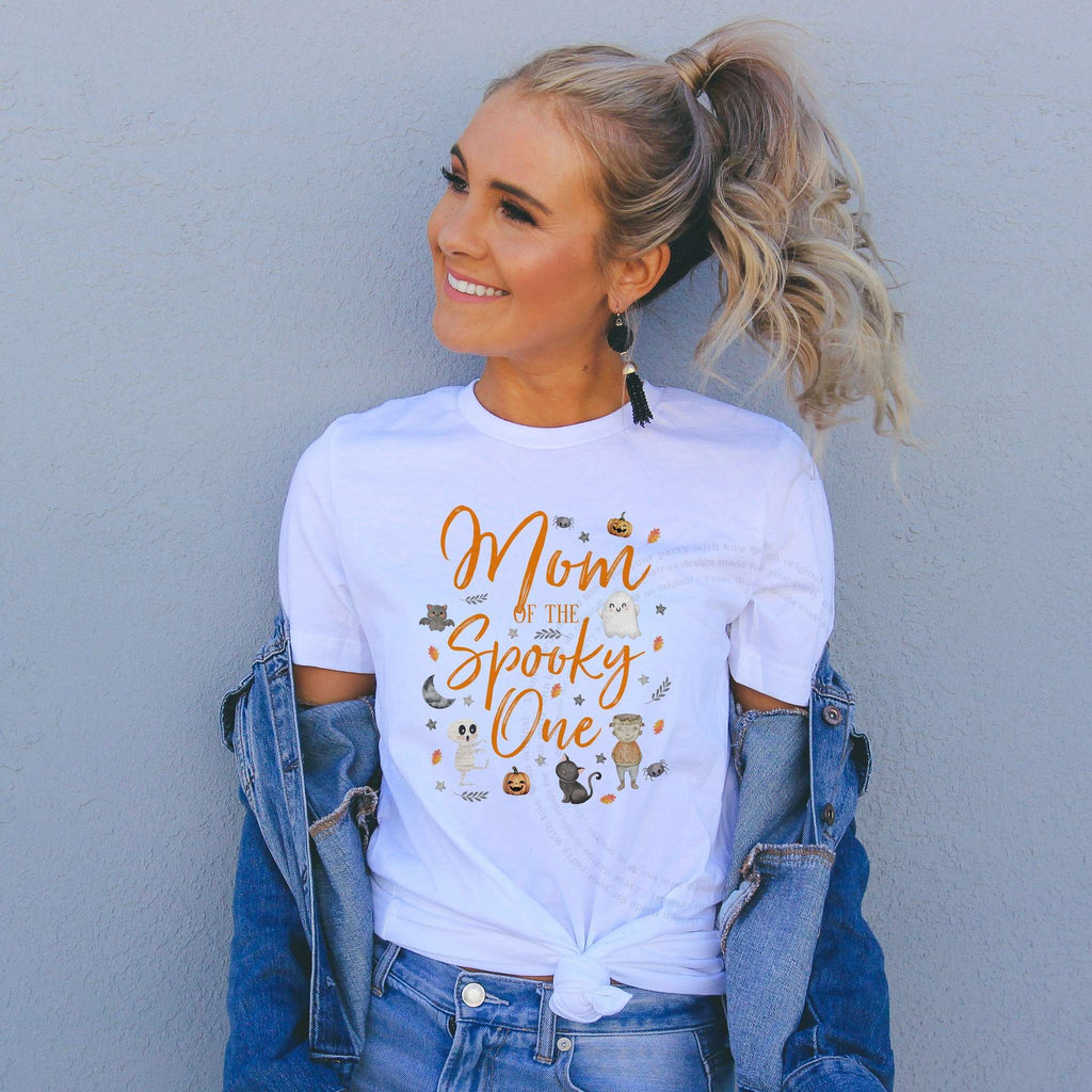 Woman in a denim jacket wearing a Mom of the Spooky One shirt with a mummy, cat and ghost.