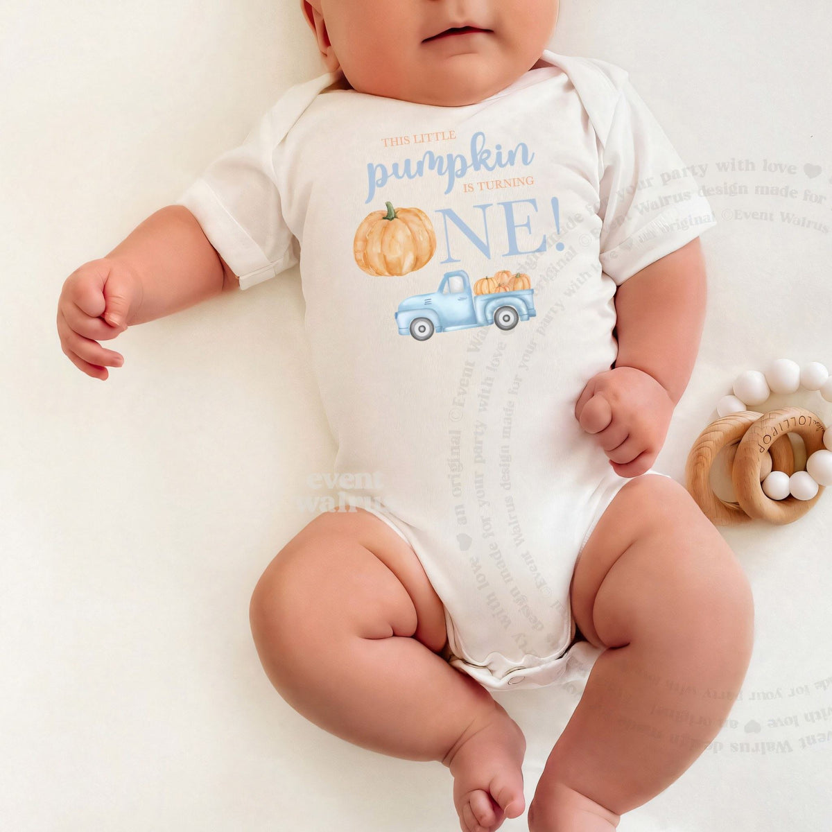 Baby lying on a white blanket in a white Little Pumpkin bodysuit with a blue truck.