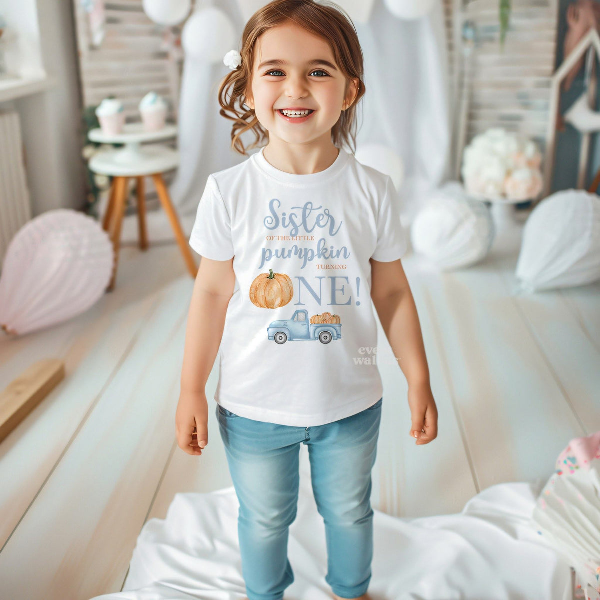 Smiling toddler girl in a white Sister of the Little Pumpkin tee in a bright party room.