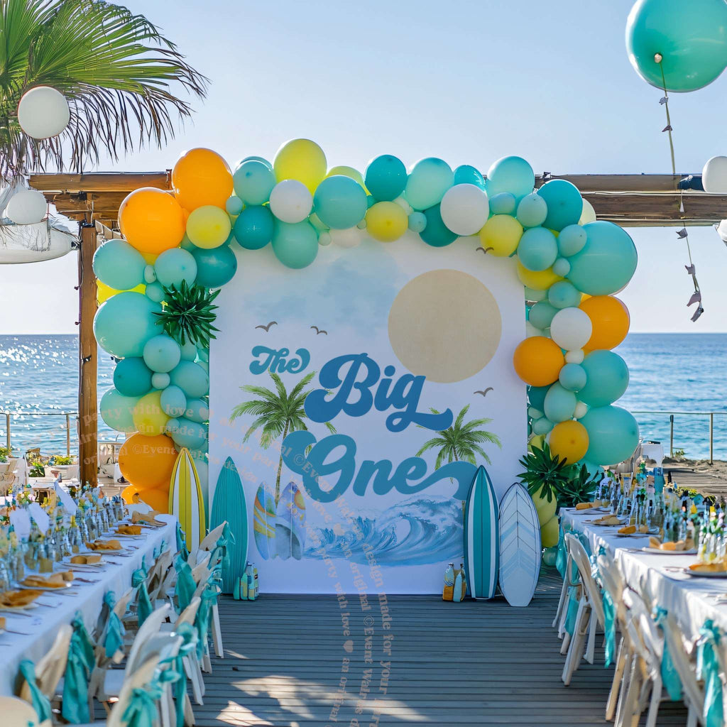The Big One backdrop with a sun and palms behind long party tables by the ocean.