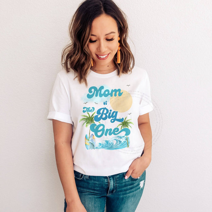 Woman in a white Mom of The Big One tee with palm trees, a sun and a blue wave.