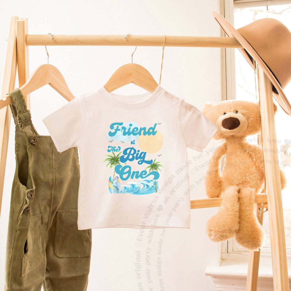 White toddler Friend of The Big One shirt on a hanger beside a teddy bear.
