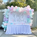 Pink, mint and lavender winter onederland backdrop behind a white dessert table outdoors.