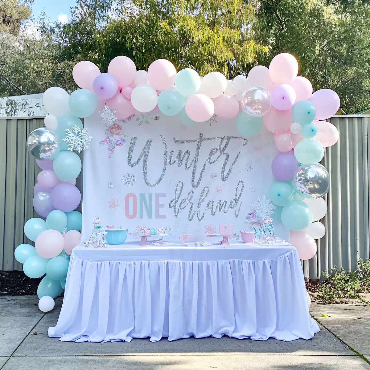 Pink, mint and lavender winter onederland backdrop behind a white dessert table outdoors.