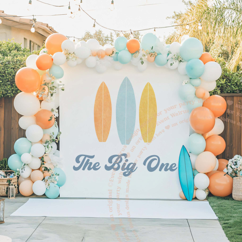 Surfboard The Big One backdrop framed by a peach and aqua balloon arch beside a blue surfboard.