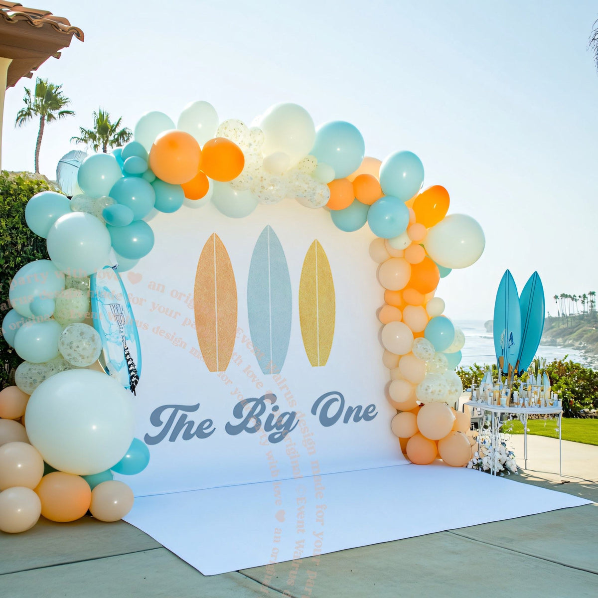 Three pastel surfboards backdrop on a lawn above the beach with a balloon arch.