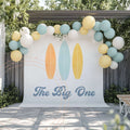 Pastel Surfboard The Big One backdrop on a patio under a blue, yellow and white balloon garland.