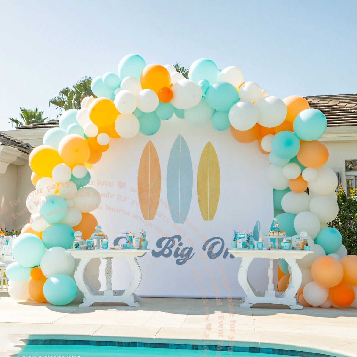 The Big One surfboard backdrop at a pool party with two white dessert tables.
