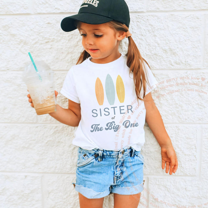 Girl in a ball cap wearing a white Sister of The Big One surfboard tee.