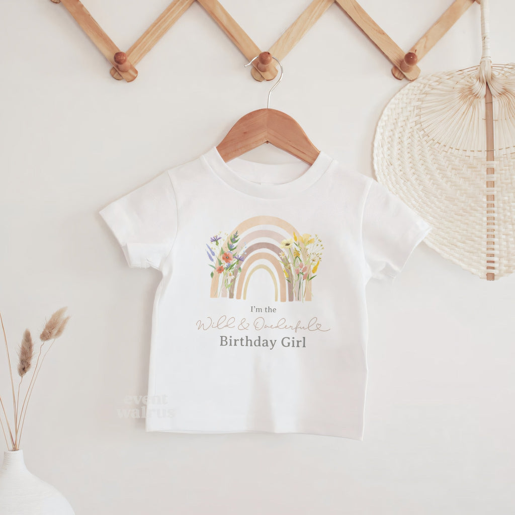 White I'm the Birthday Girl baby tee with a pastel rainbow on a wooden hanger.