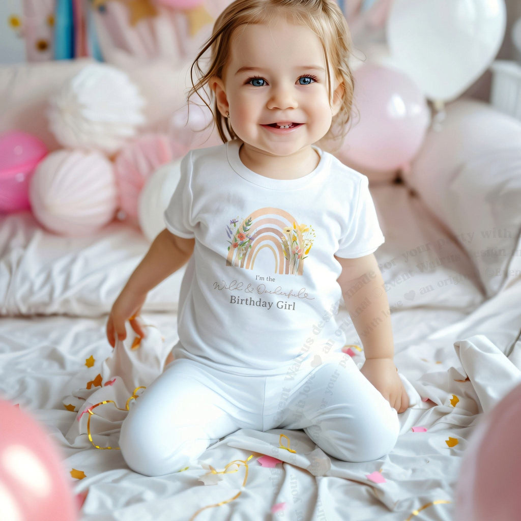 Toddler girl among pink balloons in a white I'm the Wild and Onederful Birthday Girl shirt.