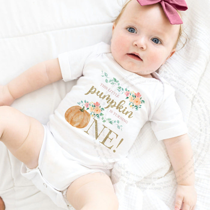 Baby girl with a pink bow lying in a white This Little Pumpkin Is Turning One bodysuit.