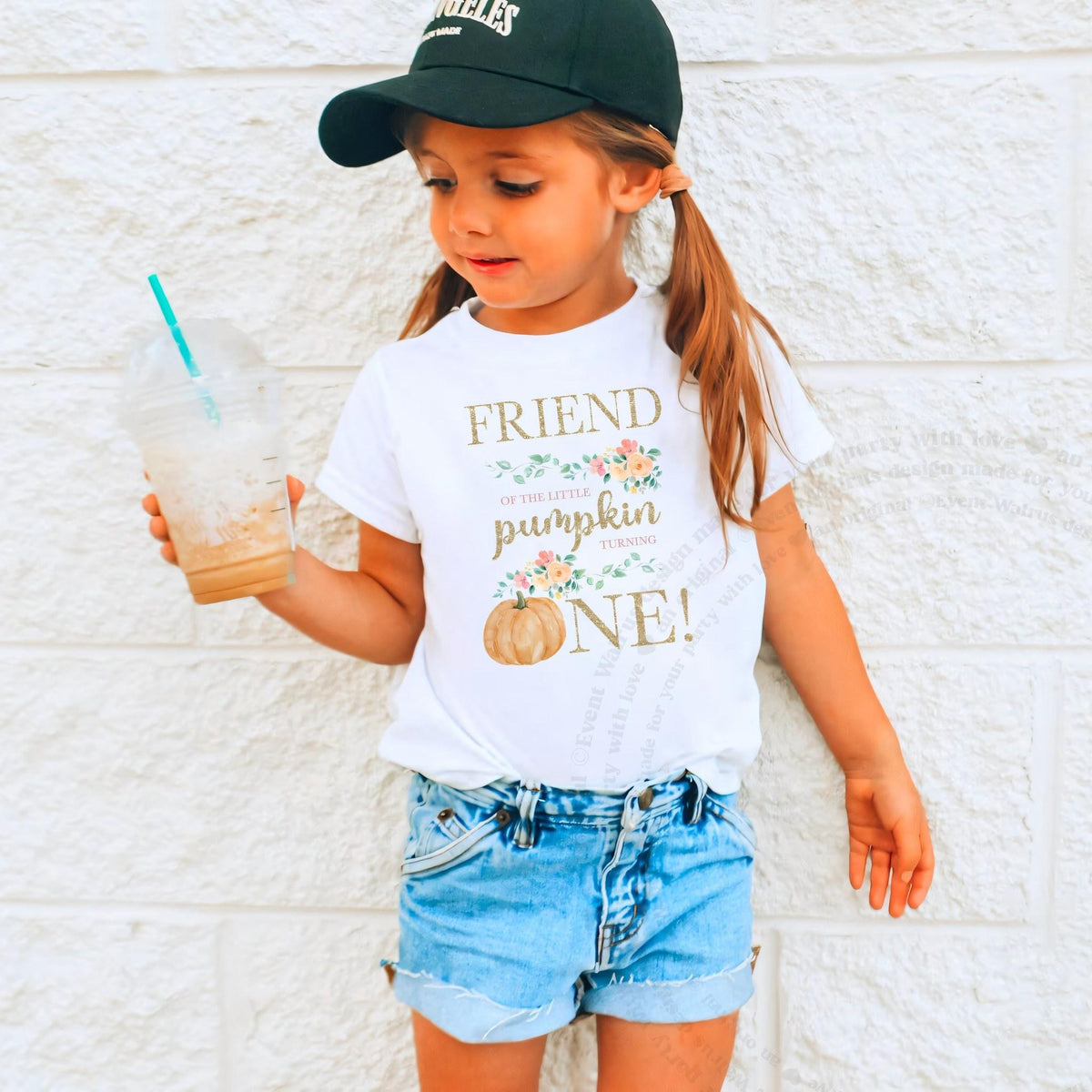 Girl in a ball cap holding a drink in a white Friend of the Little Pumpkin shirt.