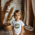 Toddler boy in a white I'm One Happy Camper tee with a wood slice ONE and pine trees.