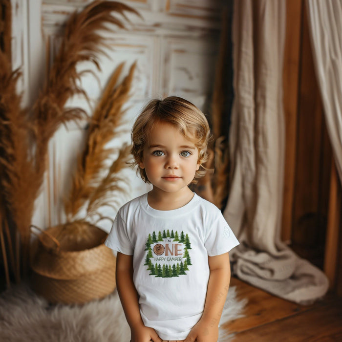 Toddler boy in a white I'm One Happy Camper tee with a wood slice ONE and pine trees.