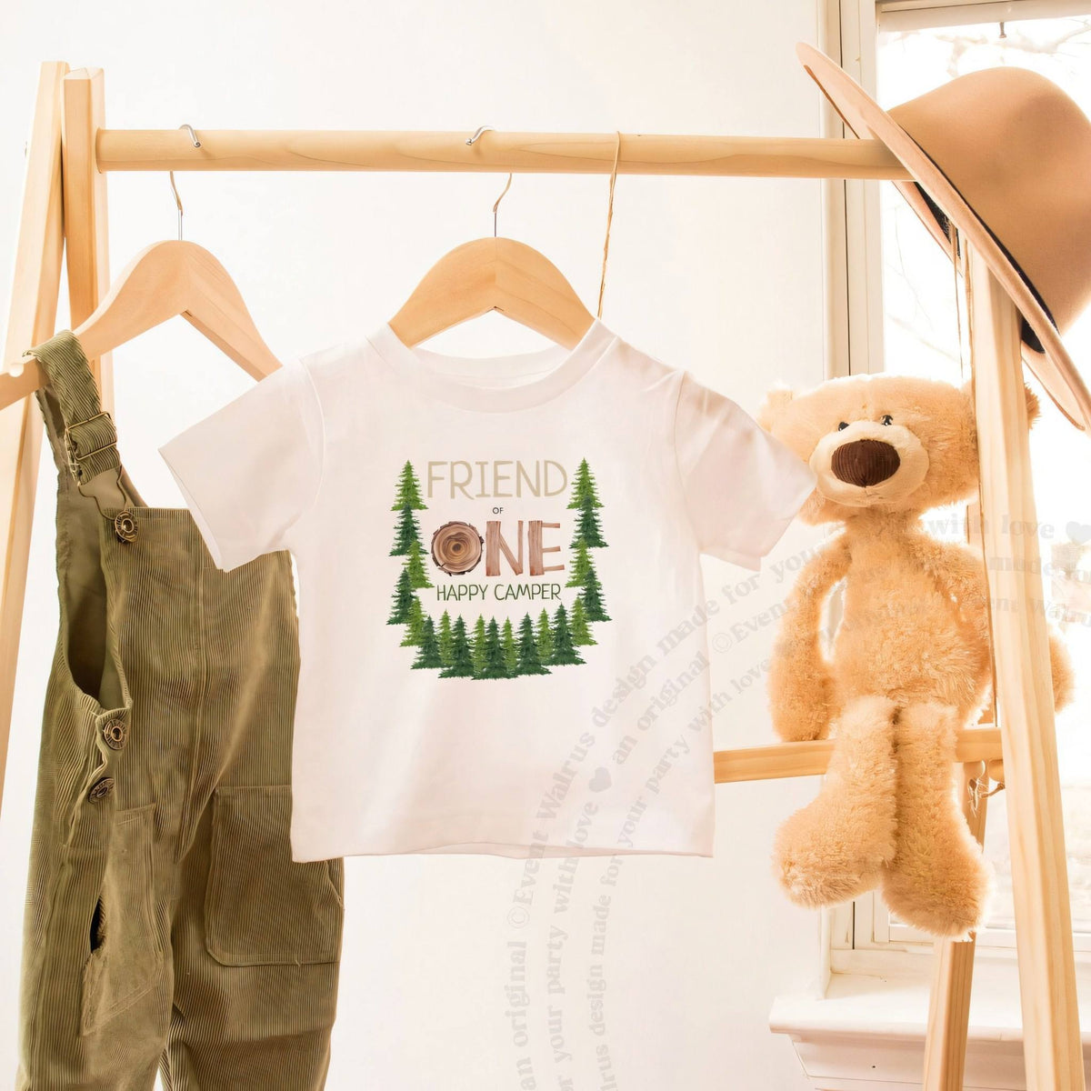 White Friend One Happy Camper toddler tee on a wooden clothes rack beside a teddy bear.