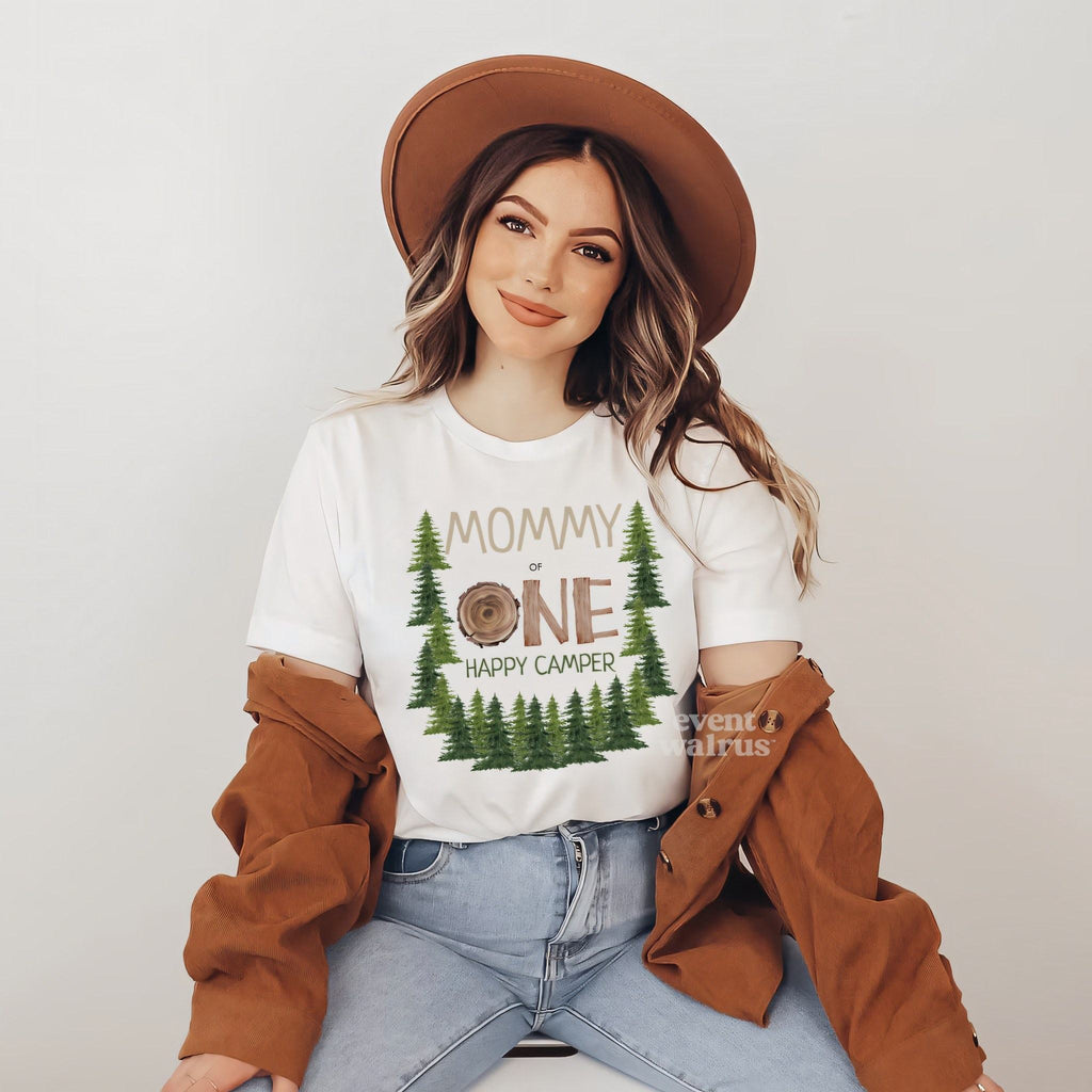 Smiling woman in a brown hat and jacket wearing a white Mommy One Happy Camper shirt with pine trees.