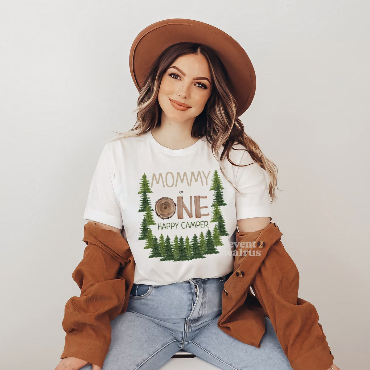 Smiling woman in a brown hat and jacket wearing a white Mommy One Happy Camper shirt with pine trees.