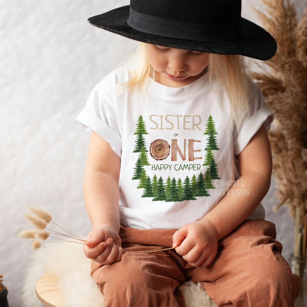 Toddler in a black hat sitting in a white Sister One Happy Camper tee.