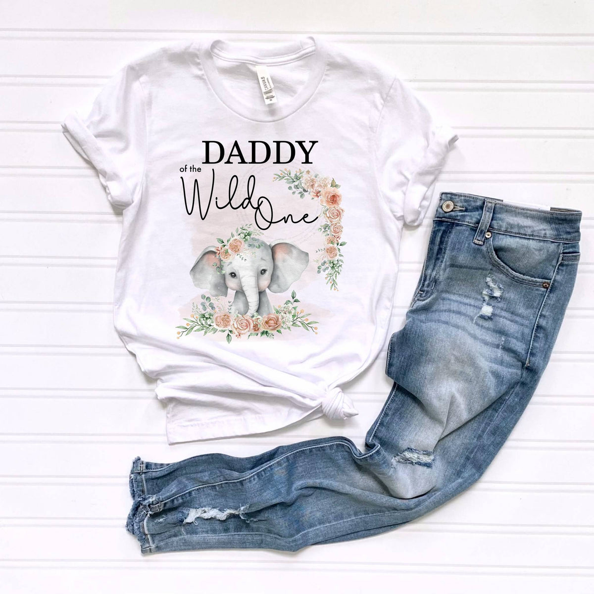 White Daddy of the Wild One adult tee with the pink elephant design, laid flat beside jeans.