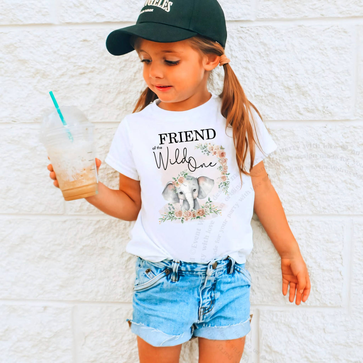 Girl in a ball cap holding a drink in a white Friend of the Wild One shirt.