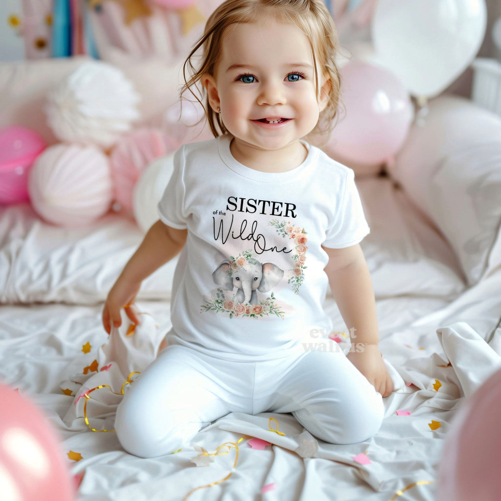 Smiling toddler girl among pink balloons in a white Sister of the Wild One elephant tee.