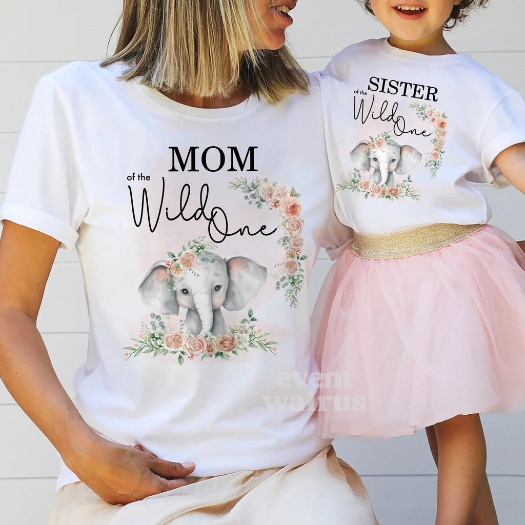 Mom holding hands with her little girl, both in white pink elephant Wild One shirts.