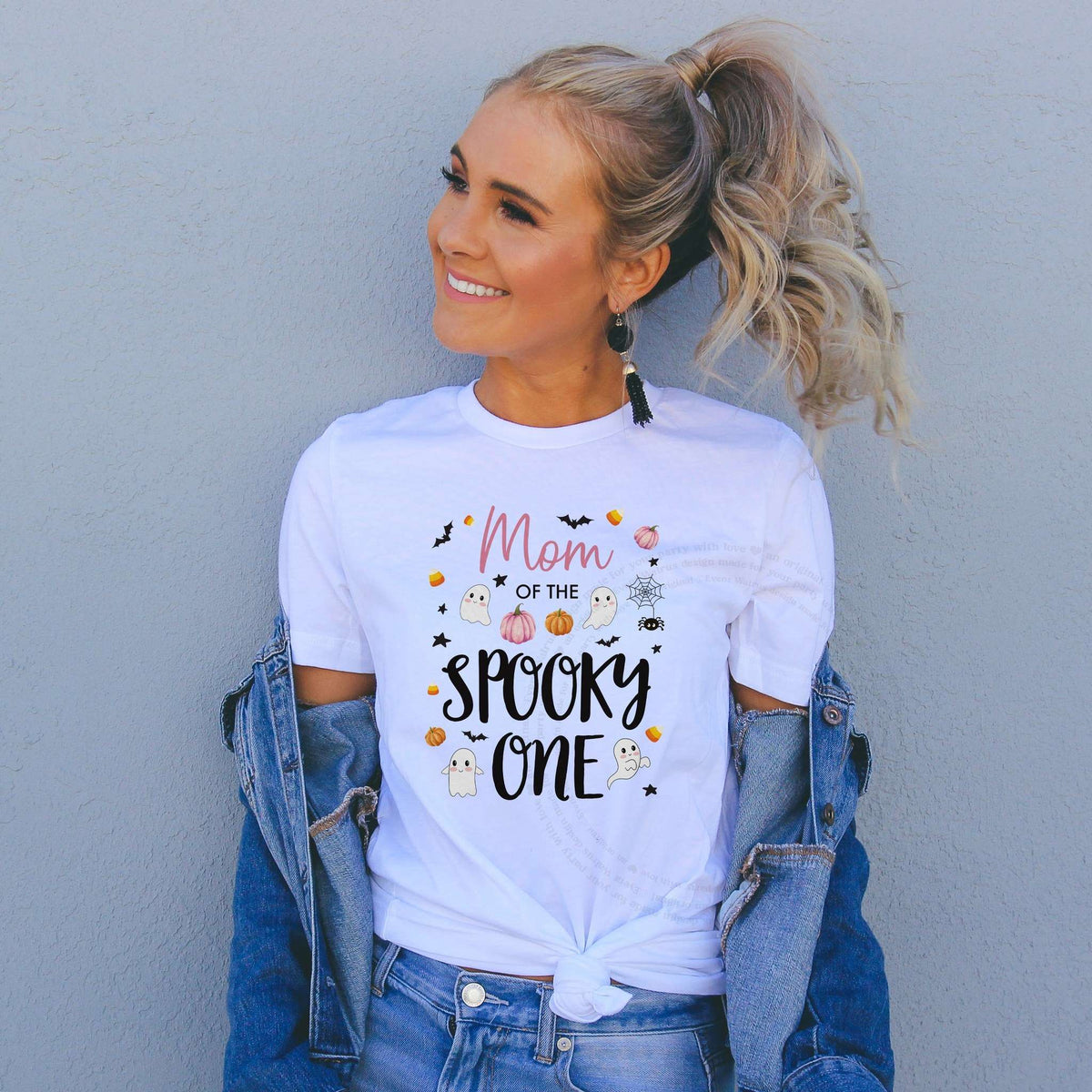 Woman in a denim jacket wearing a Mom of the Spooky One shirt with pink pumpkins and ghosts.