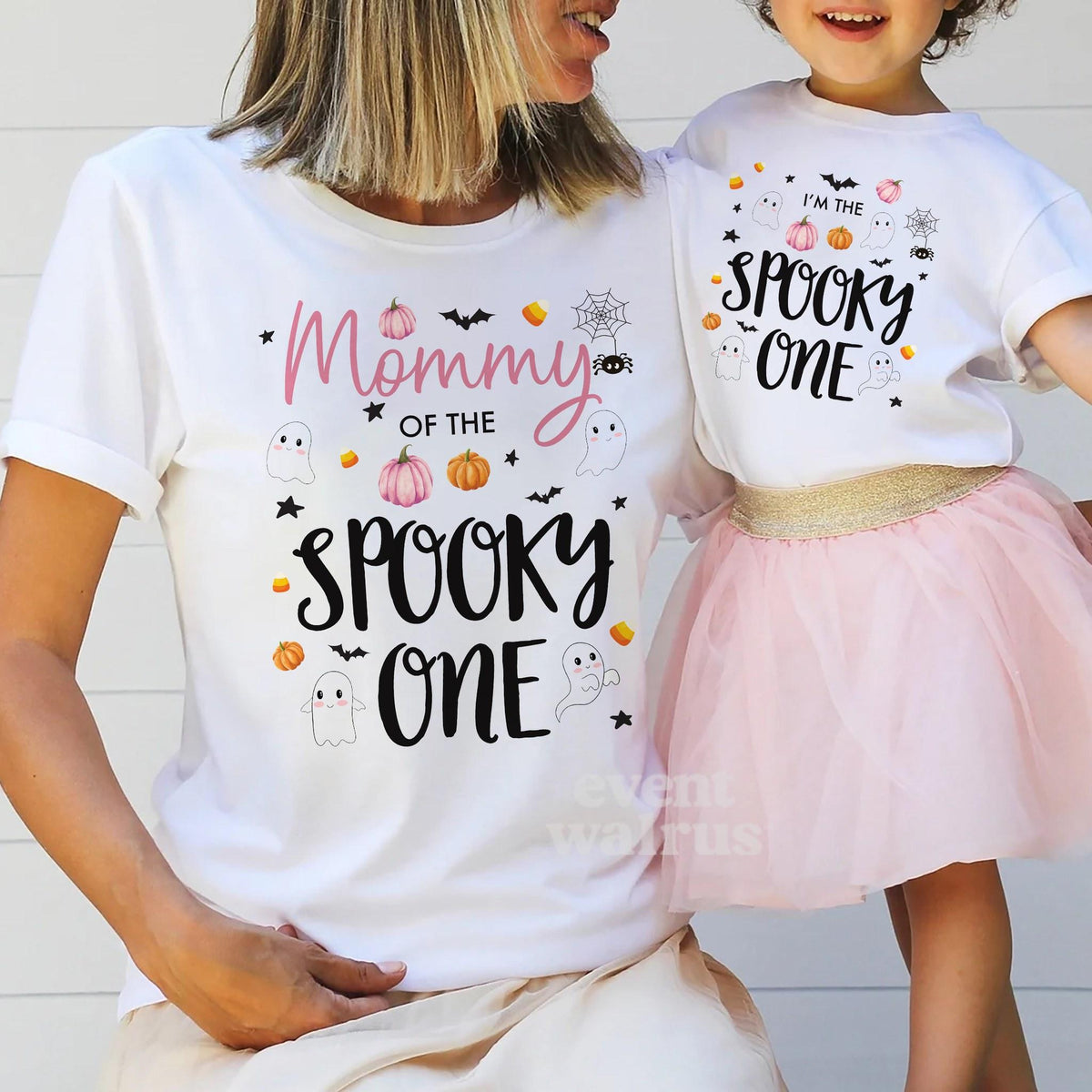 Mom holding hands with her little girl in a tutu, both in white Spooky One shirts.