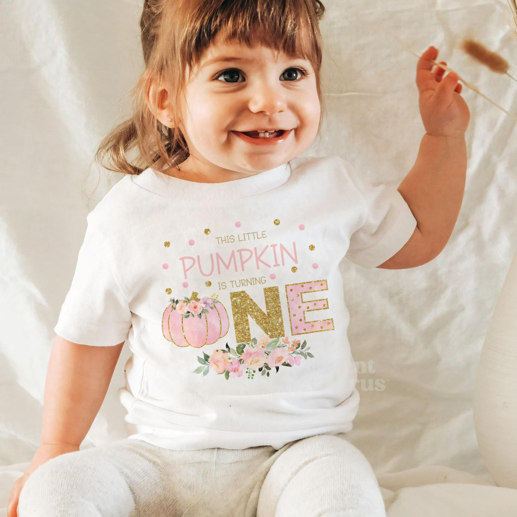 Laughing baby girl in a white This Little Pumpkin Is Turning One baby tee.