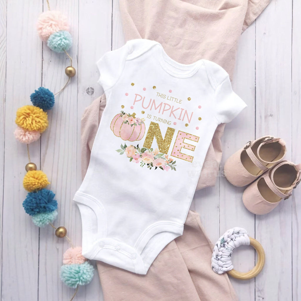 White Little Pumpkin bodysuit laid flat with pom poms, baby shoes and a blush blanket.