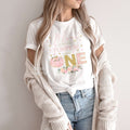 Woman in a knit cardigan wearing a white pink and gold Little Pumpkin Turning One shirt.