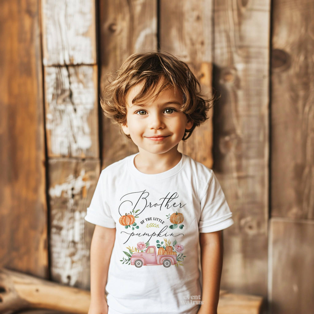 Toddler boy against a weathered wood wall in a white Brother of the Little Pumpkin tee.
