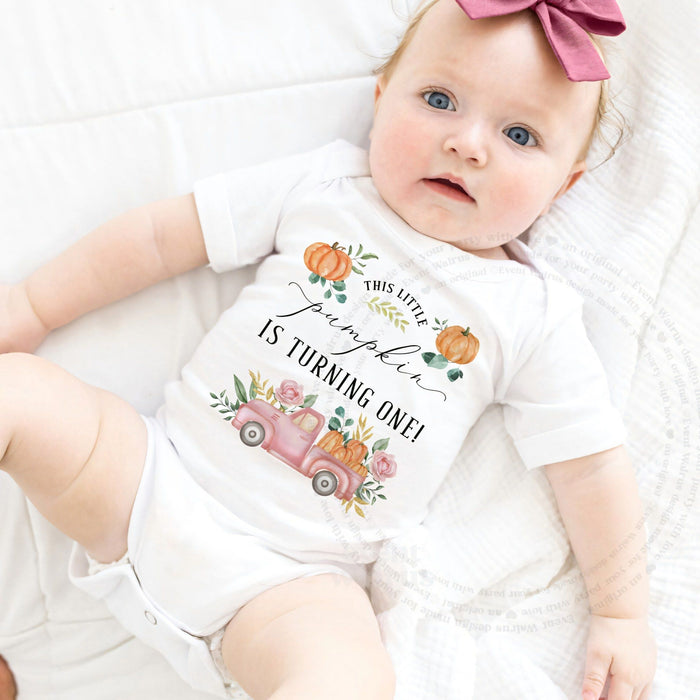 Baby girl with a pink bow lying in a white This Little Pumpkin Is Turning One bodysuit.