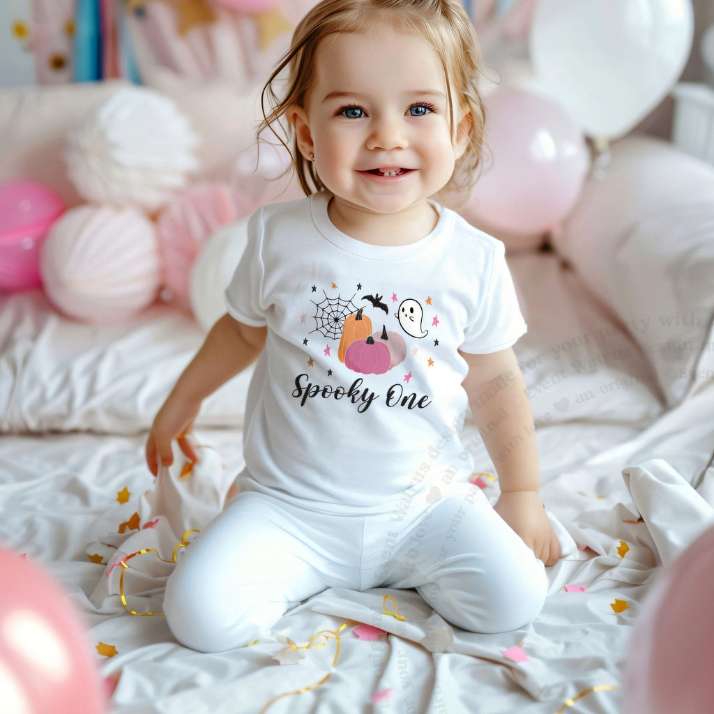 Smiling toddler girl among pink balloons in a white Spooky One tee with pink pumpkins.