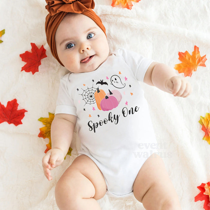 Baby girl in a rust headband lying among fall leaves in a white Spooky One bodysuit.