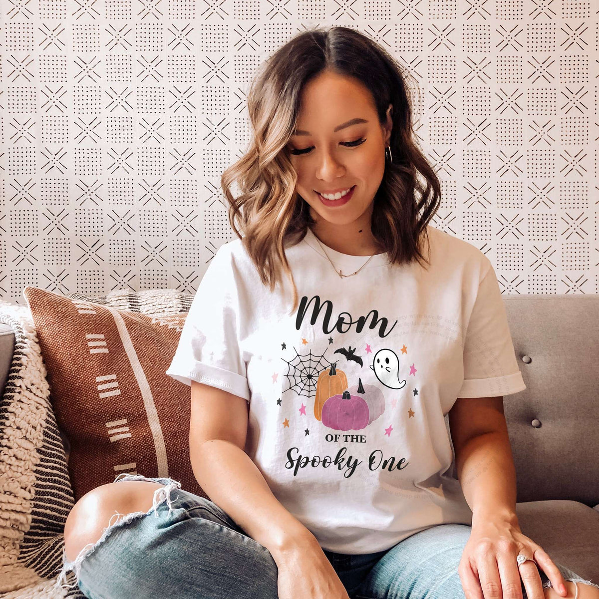 Woman sitting on a sofa with patterned pillows, wearing a white Mom of the Spooky One shirt with pink pumpkins.