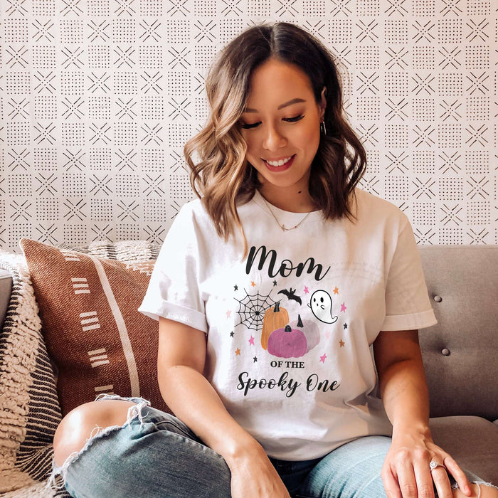 Woman sitting on a sofa with patterned pillows, wearing a white Mom of the Spooky One shirt with pink pumpkins.