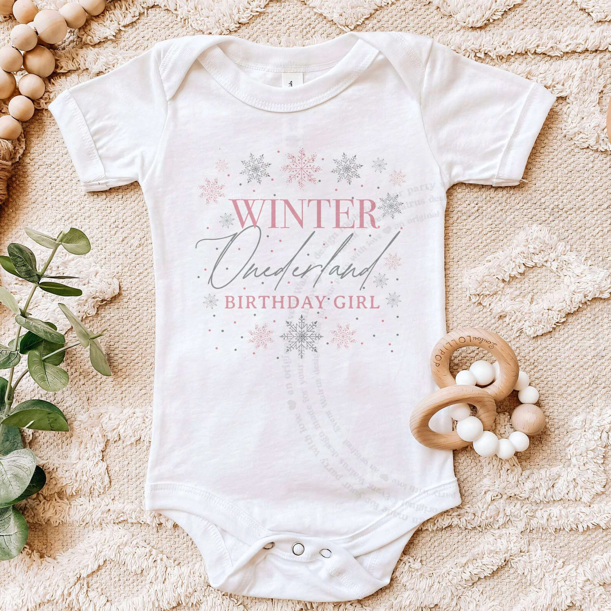 White Winter Onederland Birthday Girl bodysuit on a beige blanket with a wooden teether.