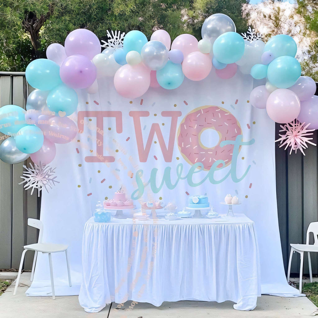 Two sweet donut backdrop behind a white dessert table with a pastel balloon arch.