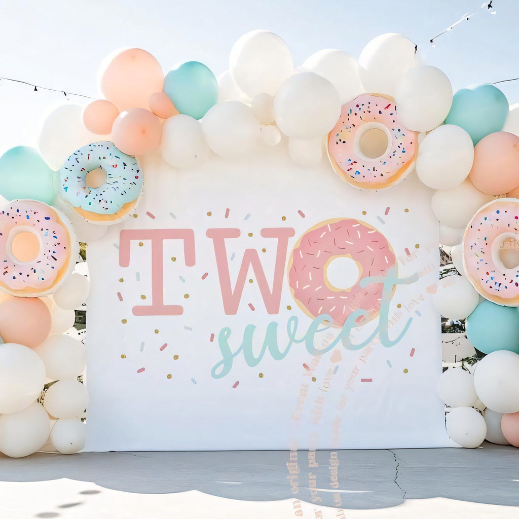 Pink sprinkle donut two sweet backdrop under a peach and mint balloon arch with donut balloons.
