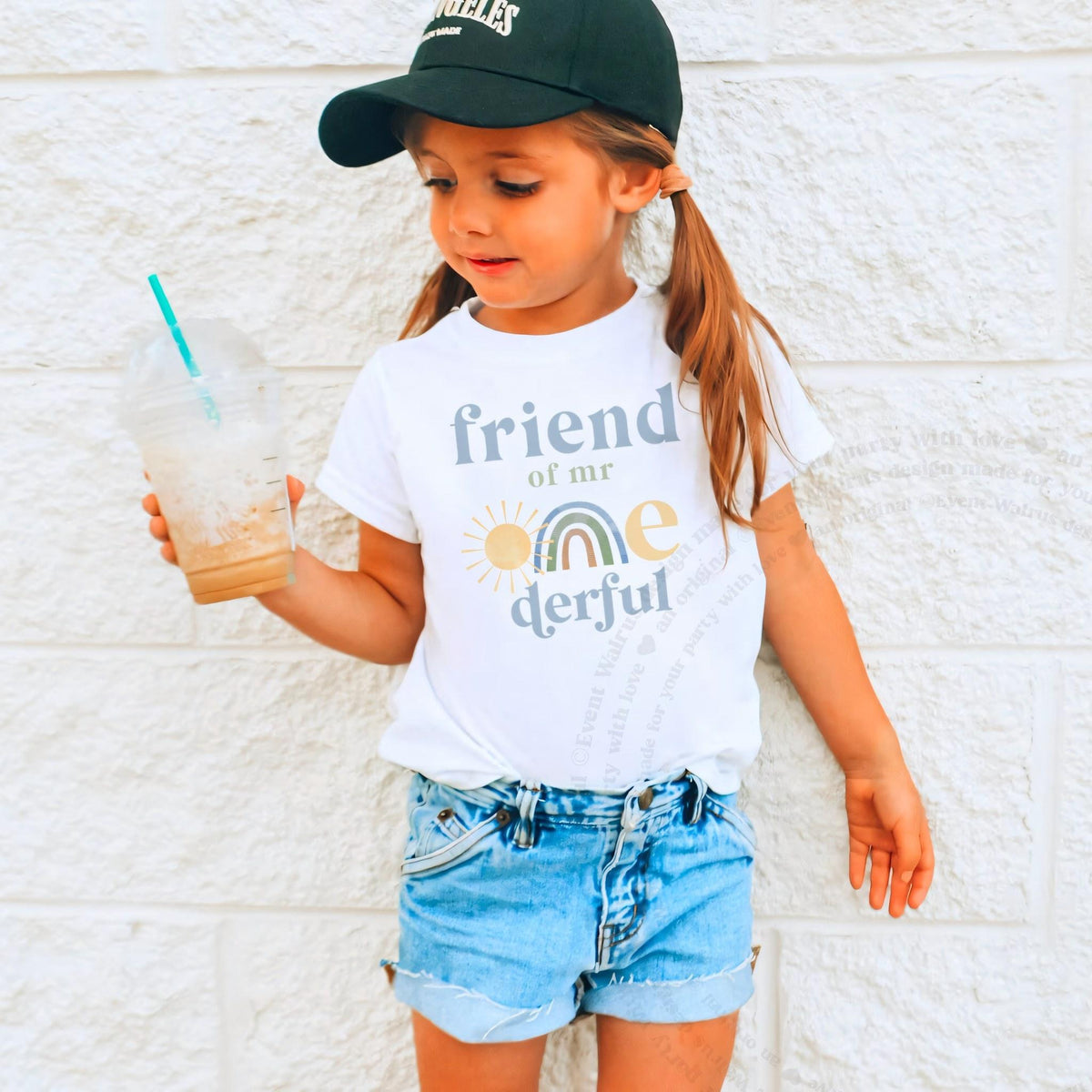 Girl in a ball cap holding a drink in a white Friend of Mr Onederful shirt.