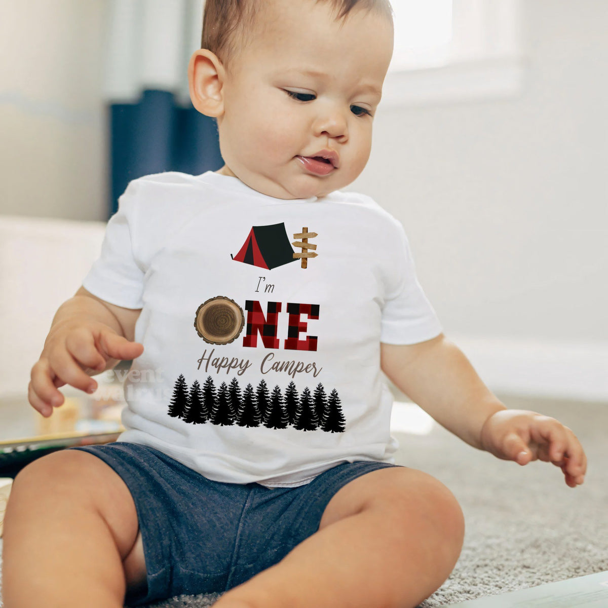 Baby boy sitting on a rug in a white I'm One Happy Camper baby tee.